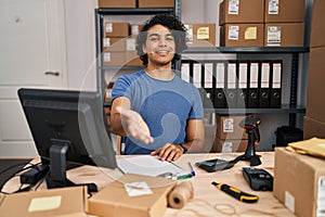 Hispanic man with curly hair working at small business ecommerce smiling cheerful offering palm hand giving assistance and