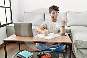 Hispanic man doing papers at home smiling with happy face looking and pointing to the side with thumb up