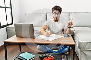 Hispanic man doing papers at home surprised pointing with finger to the side, open mouth amazed expression