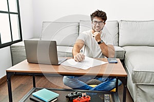 Hispanic man doing papers at home thinking worried about a question, concerned and nervous with hand on chin