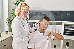 Hispanic man getting medical hearing aid at the doctor with angry face, negative sign showing dislike with thumbs down, rejection