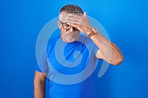 Hispanic man with grey hair standing over blue background covering eyes with hand, looking serious and sad