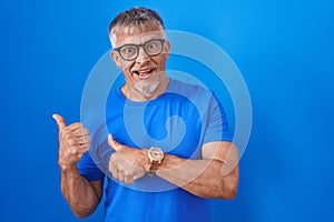 Hispanic man with grey hair standing over blue background pointing to the back behind with hand and thumbs up, smiling confident