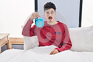 Hispanic man holding alarm clock in the bed scared and amazed with open mouth for surprise, disbelief face