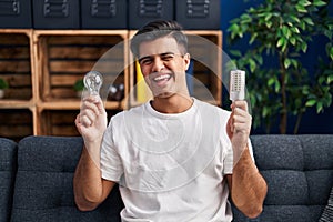 Hispanic man holding led lightbulb and incandescent bulb winking looking at the camera with sexy expression, cheerful and happy