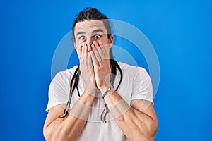 Hispanic man with long hair standing over blue background shocked covering mouth with hands for mistake