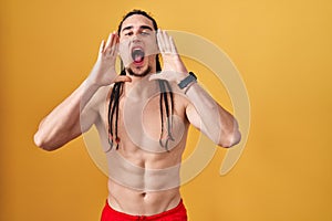 Hispanic man with long hair standing shirtless over yellow background shouting angry out loud with hands over mouth