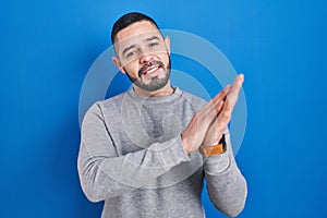 Hispanic man standing over blue background clapping and applauding happy and joyful, smiling proud hands together