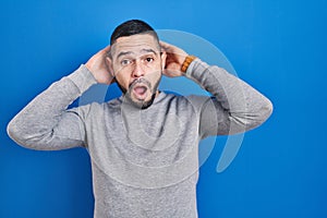 Hispanic man standing over blue background crazy and scared with hands on head, afraid and surprised of shock with open mouth
