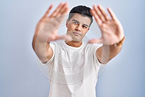 Hispanic man standing over blue background doing frame using hands palms and fingers, camera perspective