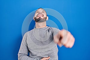 Hispanic man standing over blue background laughing at you, pointing finger to the camera with hand over body, shame expression