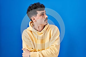 Hispanic man standing over blue background looking to the side with arms crossed convinced and confident