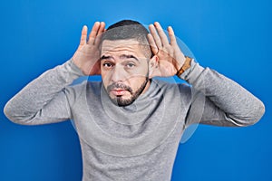 Hispanic man standing over blue background trying to hear both hands on ear gesture, curious for gossip