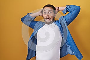 Hispanic man standing over yellow background crazy and scared with hands on head, afraid and surprised of shock with open mouth