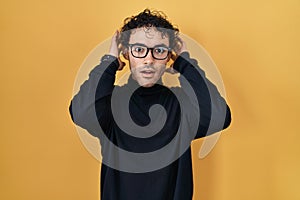 Hispanic man standing over yellow background crazy and scared with hands on head, afraid and surprised of shock with open mouth