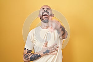 Hispanic man with tattoos standing over yellow background laughing at you, pointing finger to the camera with hand over body,