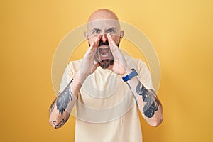 Hispanic man with tattoos standing over yellow background shouting angry out loud with hands over mouth