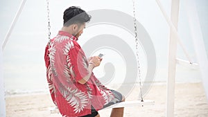 Hispanic man using smartphone on the beach outdoor with the sunlight. Attractive bearded man sitting on giant swing using smartpho