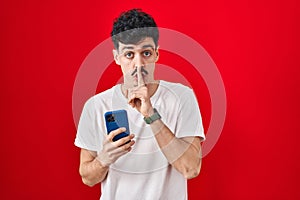 Hispanic man using smartphone over red background asking to be quiet with finger on lips