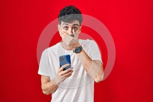 Hispanic man using smartphone over red background shocked covering mouth with hands for mistake