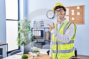 Hispanic man wearing architect hardhat smiling cheerful pointing with hand and finger up to the side