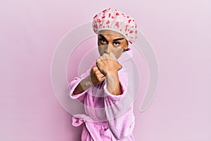 Hispanic man wearing make up wearing shower towel cap and bathrobe ready to fight with fist defense gesture, angry and upset face,