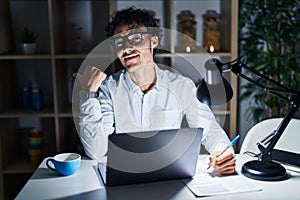 Hispanic man working at the office at night pointing to the back behind with hand and thumbs up, smiling confident