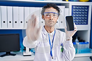 Hispanic man working at scientist laboratory showing smartphone screen with open hand doing stop sign with serious and confident