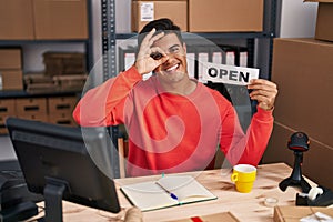 Hispanic man working at small business ecommerce holding open banner smiling happy doing ok sign with hand on eye looking through