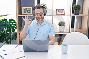 Hispanic man working using computer laptop wearing headphones doing ok sign with fingers, smiling friendly gesturing excellent