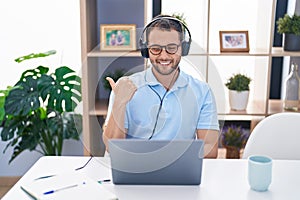 Hispanic man working using computer laptop wearing headphones pointing thumb up to the side smiling happy with open mouth