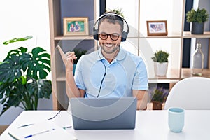 Hispanic man working using computer laptop wearing headphones smiling happy pointing with hand and finger to the side