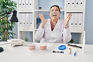 Hispanic mature dentist woman working at the clinic crazy and mad shouting and yelling with aggressive expression and arms raised