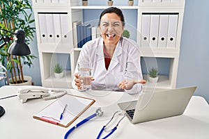 Hispanic mature doctor woman holding pill and glass of water sticking tongue out happy with funny expression