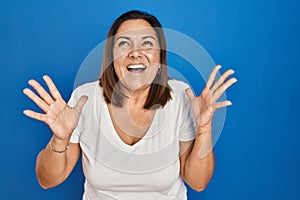 Hispanic mature woman standing over blue background crazy and mad shouting and yelling with aggressive expression and arms raised