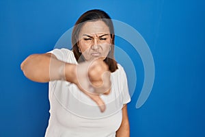 Hispanic mature woman standing over blue background looking unhappy and angry showing rejection and negative with thumbs down