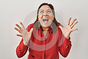 Hispanic mature woman standing over white background crazy and mad shouting and yelling with aggressive expression and arms raised