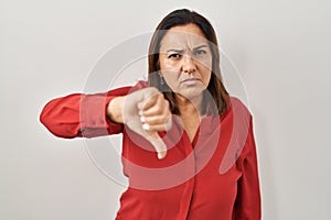 Hispanic mature woman standing over white background looking unhappy and angry showing rejection and negative with thumbs down