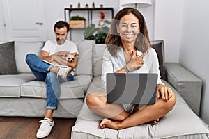 Hispanic middle age couple at home, woman using laptop cheerful with a smile of face pointing with hand and finger up to the side