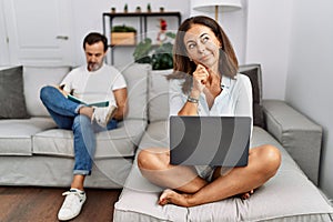 Hispanic middle age couple at home, woman using laptop looking confident at the camera smiling with crossed arms and hand raised