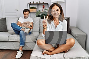 Hispanic middle age couple at home, woman using laptop smiling with happy face winking at the camera doing victory sign