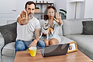 Hispanic middle age couple sitting on the sofa using computer laptop with open hand doing stop sign with serious and confident