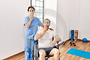 Hispanic middle age man sitting on wheelchair and nurse at rehabilitation clinic covering mouth with hand, shocked and afraid for