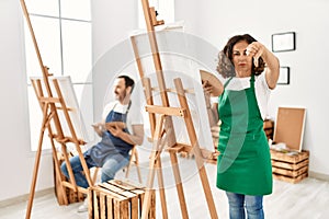 Hispanic middle age woman and mature man at art studio with angry face, negative sign showing dislike with thumbs down, rejection