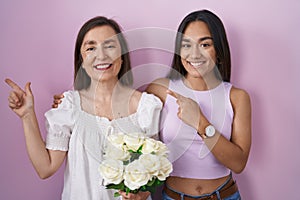 Hispanic mother and daughter holding bouquet of white flowers with a big smile on face, pointing with hand finger to the side