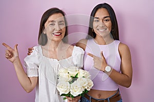 Hispanic mother and daughter holding bouquet of white flowers cheerful with a smile on face pointing with hand and finger up to