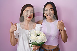 Hispanic mother and daughter holding bouquet of white flowers doing happy thumbs up gesture with hand