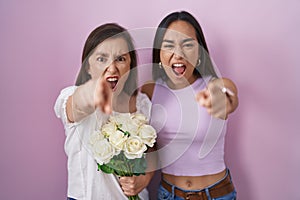 Hispanic mother and daughter holding bouquet of white flowers pointing displeased and frustrated to the camera, angry and furious