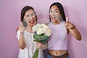 Hispanic mother and daughter holding bouquet of white flowers pointing fingers to camera with happy and funny face