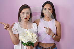 Hispanic mother and daughter holding bouquet of white flowers pointing with hand finger to the side showing advertisement, serious